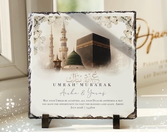 Könnte beinhalten: Eine quadratische Schieferplatte mit rauen, abgeplatzten Kanten und einem Aufdruck der Kaaba sowie der Prophetenmoschee. Sie enthält den Text "UMRAH MUBARAK", die Namen "Aisha & Yusuf" und ein Gebet für Juli 2026 auf weißem Hintergrund.