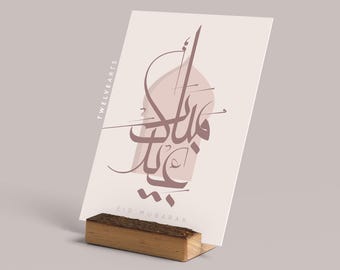 May include: A white card with a pink and brown calligraphy design that says "Eid Mubarak" in Arabic. The card is on a wooden stand and has the text "TWELVE ARTS" on the left side.