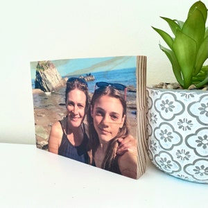 May include: A wooden block featuring a color photograph of two people on a beach. The photo is mounted on a thick, light-colored wood block, ideal for display. A decorative potted plant is visible in the background.