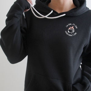 May include: Black hooded sweatshirt with a front pocket and white drawstring. The sweatshirt features a small graphic on the chest that reads "I'm fine, it's fine, everything's fine" with a burning TV graphic.