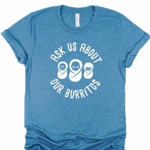 Puede incluir: Una camiseta azul con el texto "Ask Us About Our Burritos" en blanco. Tres caras de beb&eacute;s blancas est&aacute;n dentro del c&iacute;rculo de texto.
