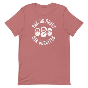 Puede incluir: Una camiseta rosa con un dise&ntilde;o circular blanco que dice "Ask Us About Our Burritos" con tres burritos beb&eacute;s dentro del c&iacute;rculo.