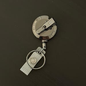 May include: A silver retractable badge reel with a metal clip. The reel has the words "YOU'RE AWESOME" and "GREAT SHIFT!" printed on it. A clear plastic strap and a metal ring are attached.