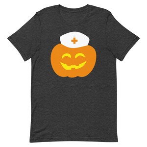 K&ouml;nnte beinhalten: Ein dunkelgraues T-Shirt mit einem Aufdruck einer l&auml;chelnden K&uuml;rbis, die einen wei&szlig;en Krankenschwesterhut mit einem roten Kreuz tr&auml;gt.