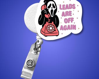 May include: A white retractable badge reel with a metal clip. The reel features a cartoon image of the Ghostface character from the Scream movies holding a red telephone. Text on the image reads "LEADS ARE OFF AGAIN."