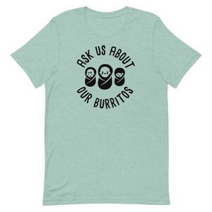 Puede incluir: Una camiseta azul claro con un gr&aacute;fico circular negro que dice "Ask Us About Our Burritos" con tres caras de beb&eacute;s dentro del c&iacute;rculo.