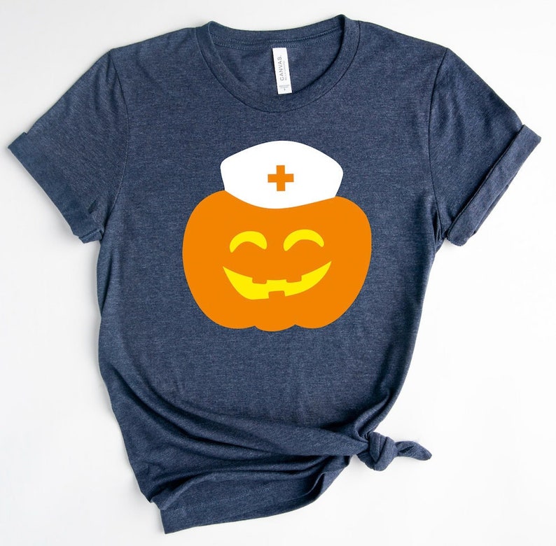 K&ouml;nnte beinhalten: Ein dunkelblaues T-Shirt mit einem orangefarbenen K&uuml;rbis, der einen wei&szlig;en Krankenschwesterhut mit einem roten Kreuz tr&auml;gt. Der K&uuml;rbis hat ein gelbes, l&auml;chelndes Gesicht.