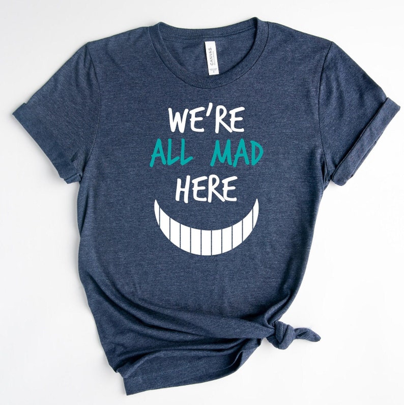 May include: A navy blue t-shirt with white text that reads "We're All Mad Here" and a white Cheshire Cat grin with teal teeth.