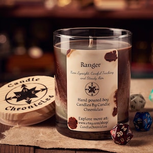 May include: A brown glass candle jar with a label that reads "Ranger" and "Keen Eyesight, Careful Tracking and Steady Aim". The candle is surrounded by dice and a wooden lid with the words "Candle Chronicles" on it.