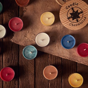May include: A collection of colorful tea light candles in various shades, including red, blue, and yellow, arranged on a wooden surface. A wooden lid with the text "Candle Chronicles" is also present. The candles are in clear holders.