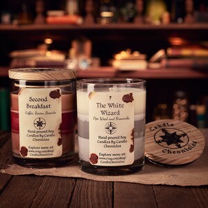May include: Two glass jar candles with wooden lids and labels. One candle is labeled "Second Breakfast" and the other "The White Wizard." The labels include the words "Hand poured Soy Candles By Candle Chronicles."