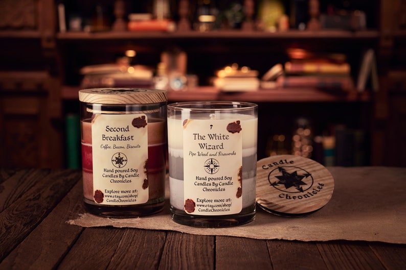 May include: Two glass jar candles with wooden lids and labels. One candle is labeled "Second Breakfast" and the other "The White Wizard." The labels include the words "Hand poured Soy Candles By Candle Chronicles."
