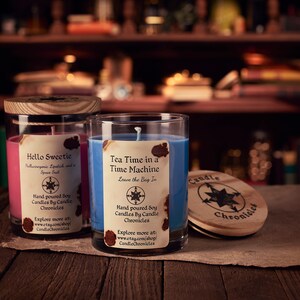 May include: Two glass jar candles with wooden lids. One candle is pink with the text "Hello Sweetie", the other is blue with the text "Tea Time in a Time Machine". A wooden lid with the text "Candle Chronicles" is also visible.