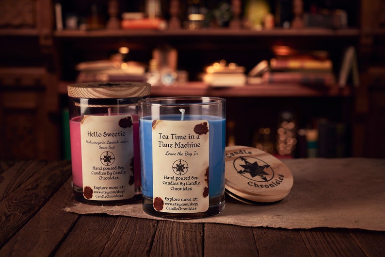 May include: Two glass jar candles with wooden lids. One candle is pink with the text "Hello Sweetie", the other is blue with the text "Tea Time in a Time Machine". A wooden lid with the text "Candle Chronicles" is also visible.