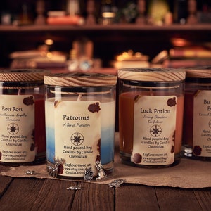 May include: Four glass jar candles with wooden lids, each labeled with a different name: "Ron Ron," "Patronus," "Luck Potion," and "Buttery Brew." The candles are on a wooden surface, with small charms scattered around.