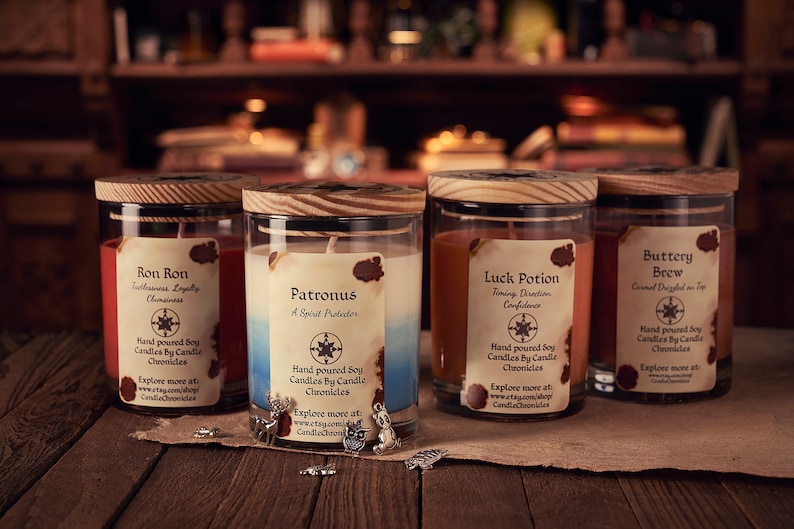 May include: Four glass jar candles with wooden lids, each labeled with a different name: "Ron Ron," "Patronus," "Luck Potion," and "Buttery Brew." The candles are on a wooden surface, with small charms scattered around.