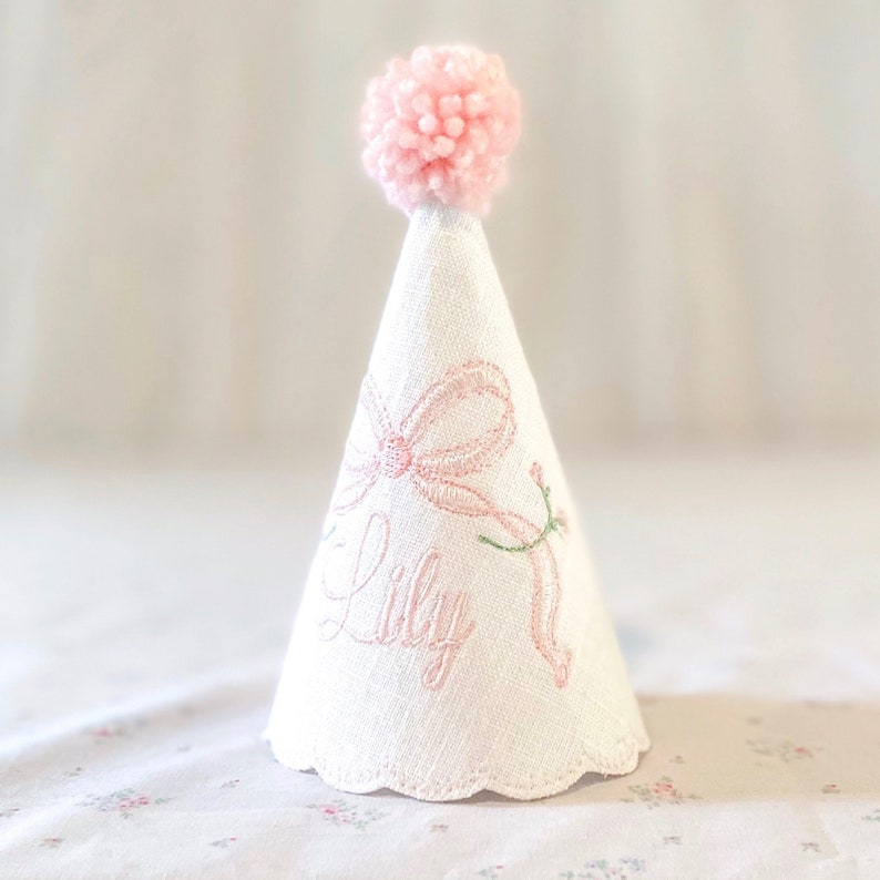 May include: A white cone-shaped party hat featuring a pink pom-pom on top. The hat is embroidered with a pink bow and the name Lily in cursive script, accented by a small green stem detail. The bottom edge of the hat has a scalloped finish.
