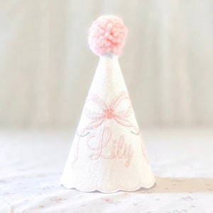 May include: A white cone-shaped party hat featuring a soft pink pom-pom at the top. The hat is decorated with an embroidered pink bow and the name Lily in cursive script. The bottom edge of the hat has a scalloped finish with delicate stitching.