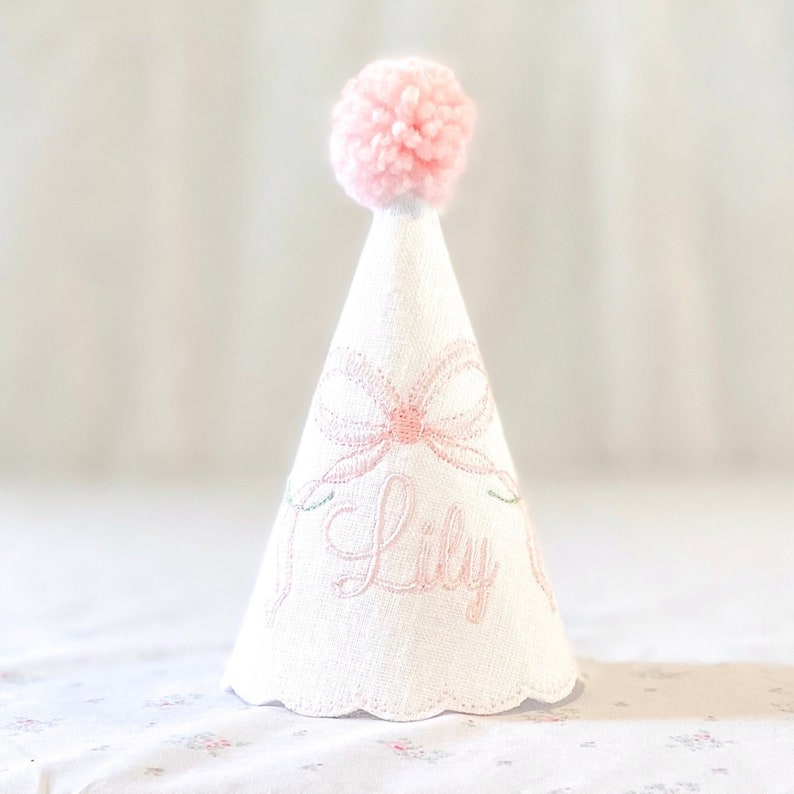 May include: A white cone-shaped party hat featuring a soft pink pom-pom at the top. The hat is decorated with an embroidered pink bow and the name Lily in cursive script. The bottom edge of the hat has a scalloped finish with delicate stitching.