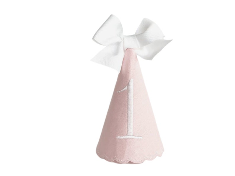 May include: A light pink party hat with a white bow at the top. The number "1" is embroidered in white on the front of the cone-shaped hat. The bottom edge of the hat has a scalloped design.