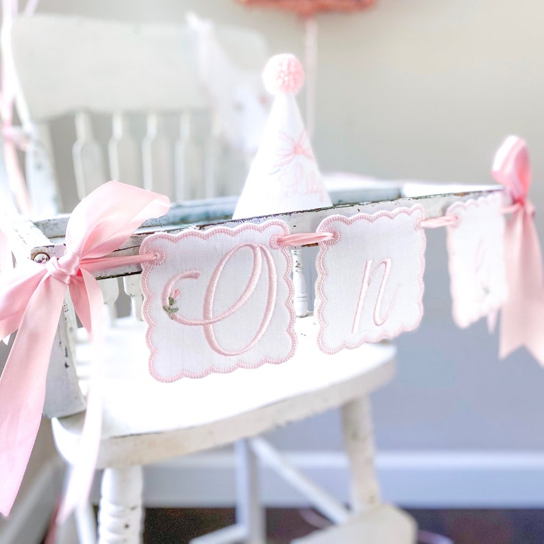 May include: A white fabric banner with scalloped edges and pink embroidered lettering spelling out the word one. The banner features light pink satin ribbon ties on each end and is accented with small embroidered floral details on the letters.