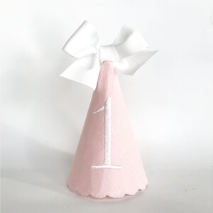 May include: A pink party hat with a white embroidered number 1 and a white bow on top. The hat has a scalloped edge and is designed for a first birthday celebration.