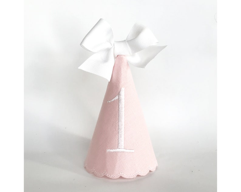 May include: A pink party hat with a white embroidered number 1 and a white bow on top. The hat has a scalloped edge and is designed for a first birthday celebration.