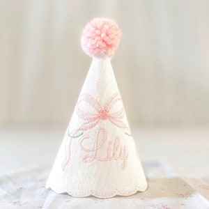 May include: A white cone-shaped party hat featuring a pink embroidered bow and the name Lily in pink script. The hat is topped with a fluffy pink pom-pom and has a scalloped bottom edge. The fabric has a visible woven texture throughout the surface.