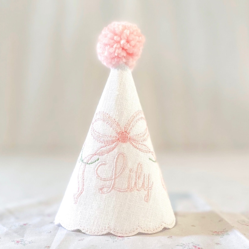 May include: A white cone-shaped party hat featuring a pink embroidered bow and the name Lily in pink script. The hat is topped with a fluffy pink pom-pom and has a scalloped bottom edge. The fabric has a visible woven texture throughout the surface.