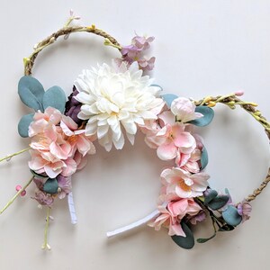 Puede incluir: Una diadema floral con un gran crisantemo blanco, flores rosas y moradas y hojas verdes. La diadema tiene un diseño circular tejido. Las flores están agrupadas alrededor de la diadema.
