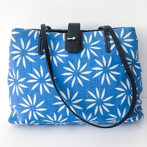 May include: A blue canvas tote bag with white floral print and black leather straps and handle.