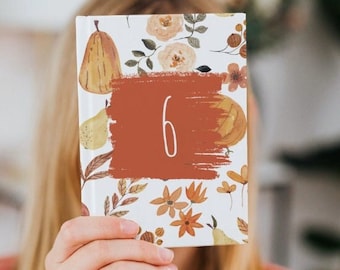May include: A white journal with a floral pattern featuring pears, leaves, and flowers in shades of orange, yellow, and brown. The number "6" is written in white on a red background.