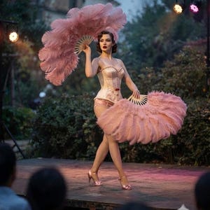 May include: A performer wearing a pale pink corset and matching bottoms, holding two large, fluffy pink feather fans. The outfit features a structured bodice with a small red accent at the waist, paired with light-colored stockings and pink heels.