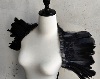 Könnte beinhalten: Schwarzes Federschulterstück auf einem weißen Mannequin. Die Federn sind in einer geschichteten, flügelartigen Form angeordnet.