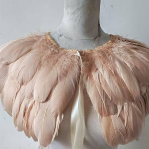 May include: A blush-toned feather capelet with a layered design. It is fastened with a cream satin ribbon. The feathers are arranged in a circular pattern, creating a soft, textured look. The capelet is displayed on a mannequin.