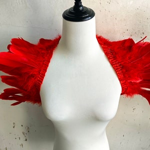 Puede incluir: Pieza de hombro de plumas rojas sobre un maniquí blanco. Las plumas están dispuestas en un diseño en capas y en cascada.