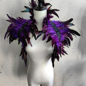 以下が含まれることがあります： 白いマネキンに紫と黒の羽根の肩飾り。羽根は重ねて滝のように配置されています。