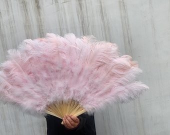 May include: A large, open hand fan with a light pink feather design. The feathers are a soft, fluffy texture, and the fan's base is made of light-colored wood. The fan is held against a textured, light gray background.