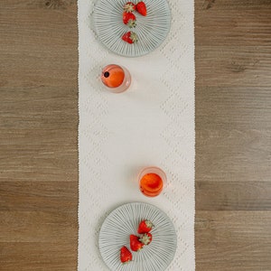K&ouml;nnte beinhalten: Ein wei&szlig;er Tischl&auml;ufer mit Rautenmuster, zwei graue Teller mit Erdbeeren und zwei Gl&auml;ser mit rosa Fl&uuml;ssigkeit. Der Tischl&auml;ufer liegt auf einem Holztisch.