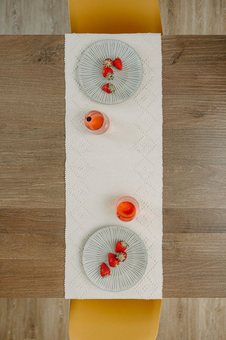 K&ouml;nnte beinhalten: Ein wei&szlig;er Tischl&auml;ufer mit Rautenmuster, zwei graue Teller mit Erdbeeren und zwei Gl&auml;ser mit rosa Fl&uuml;ssigkeit. Der Tischl&auml;ufer liegt auf einem Holztisch.