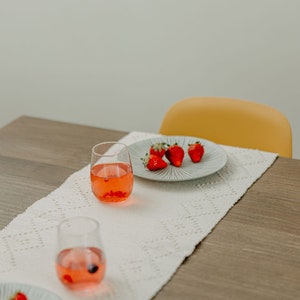 K&ouml;nnte beinhalten: Ein wei&szlig;er Tischl&auml;ufer mit einem zarten Muster liegt auf einem Holztisch. Zwei klare Gl&auml;ser gef&uuml;llt mit einer rosa Fl&uuml;ssigkeit und ein wei&szlig;er Teller mit roten Erdbeeren stehen auf dem L&auml;ufer. Ein gelber Stuhl befindet sich im Hintergrund.