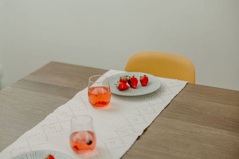 K&ouml;nnte beinhalten: Ein wei&szlig;er Tischl&auml;ufer mit einem zarten Muster liegt auf einem Holztisch. Zwei klare Gl&auml;ser gef&uuml;llt mit einer rosa Fl&uuml;ssigkeit und ein wei&szlig;er Teller mit roten Erdbeeren stehen auf dem L&auml;ufer. Ein gelber Stuhl befindet sich im Hintergrund.