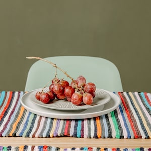 Pode incluir: Um cacho de uvas vermelhas em um prato cinza com borda branca, colocado sobre um corredor de mesa listrado colorido. O prato está em uma mesa de madeira, com uma cadeira verde claro ao fundo.