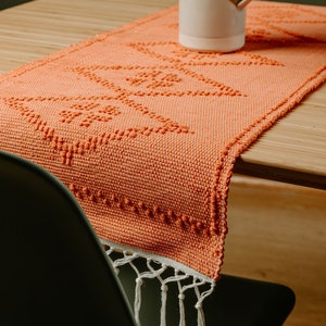 Könnte beinhalten: Orangefarbener Tischläufer mit gewebtem Muster und weißen Fransen. Der Läufer liegt auf einem Holztisch mit einer weißen Vase und getrockneten Blumen.