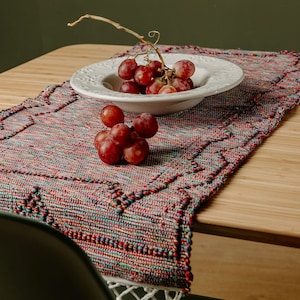Puede incluir: Un cuenco de cerámica blanca lleno de uvas rojas se encuentra sobre un camino de mesa tejido de colores con flecos blancos. El cuenco está sobre una mesa de madera.