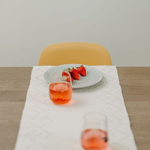 K&ouml;nnte beinhalten: Ein wei&szlig;er Tischl&auml;ufer mit einem wei&szlig;en H&auml;kelmuster, ein Teller mit drei roten Erdbeeren und zwei klare Gl&auml;ser gef&uuml;llt mit einer rosa Fl&uuml;ssigkeit.