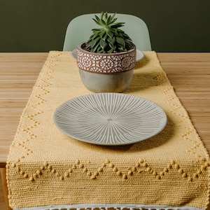 Pode incluir: Um corredor de mesa amarelo com franjas brancas e um padrão geométrico. Um prato branco e uma suculenta em vaso estão em cima do corredor de mesa.