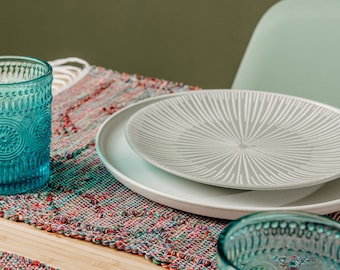 May include: Two teal blue glasses with a textured pattern sit on a colorful woven placemat with a white plate and a gray plate with a sunburst pattern on top.