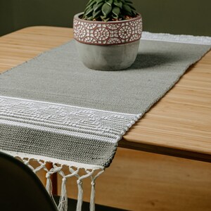May include: A gray woven table runner with white trim and tassels. The runner is on a wooden table with a small potted succulent plant in the center.