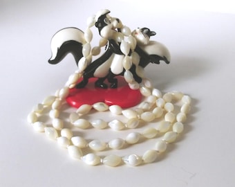 May include: A vintage figurine of two skunks in black and white, standing on a red heart-shaped base, with a long, off-white beaded necklace draped around them. The necklace is made of oval-shaped beads.
